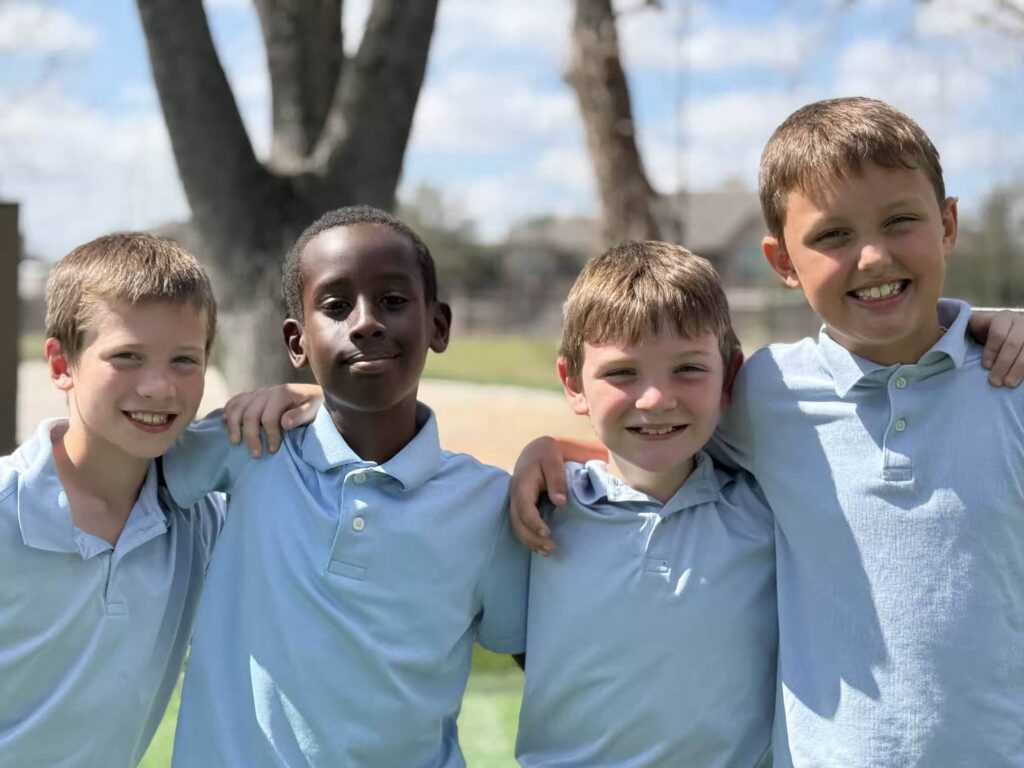 Four young boys in light blue shirts, smiling and posing together outdoors.