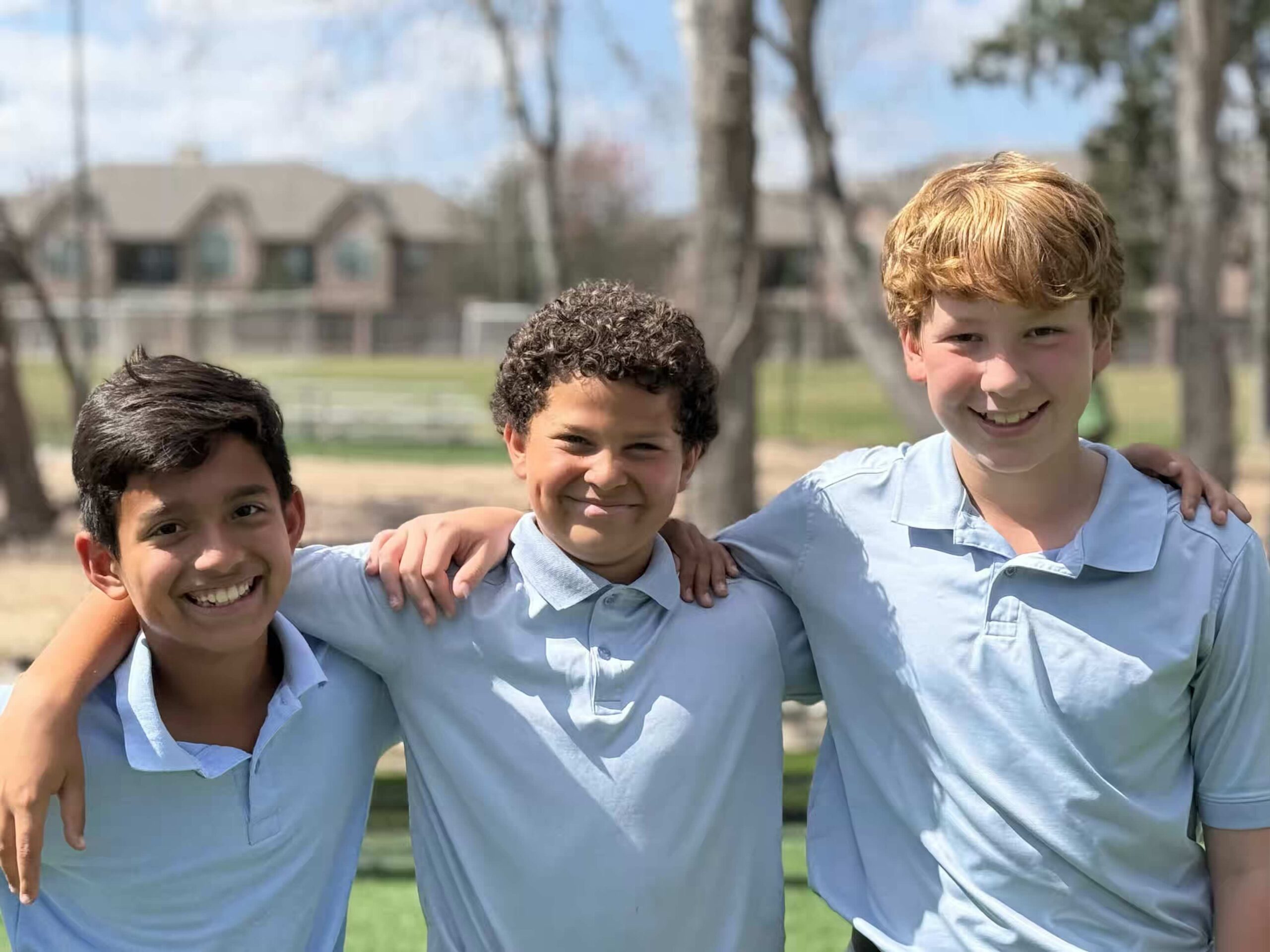 Three smiling young boys in light blue polo shirts with arms around each other outdoors.