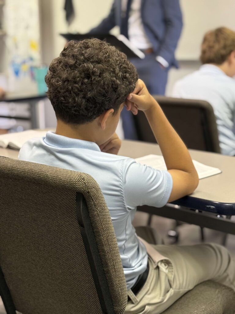 Student in light blue polo shirt and khaki pants sits at desk, looking towards teacher.
