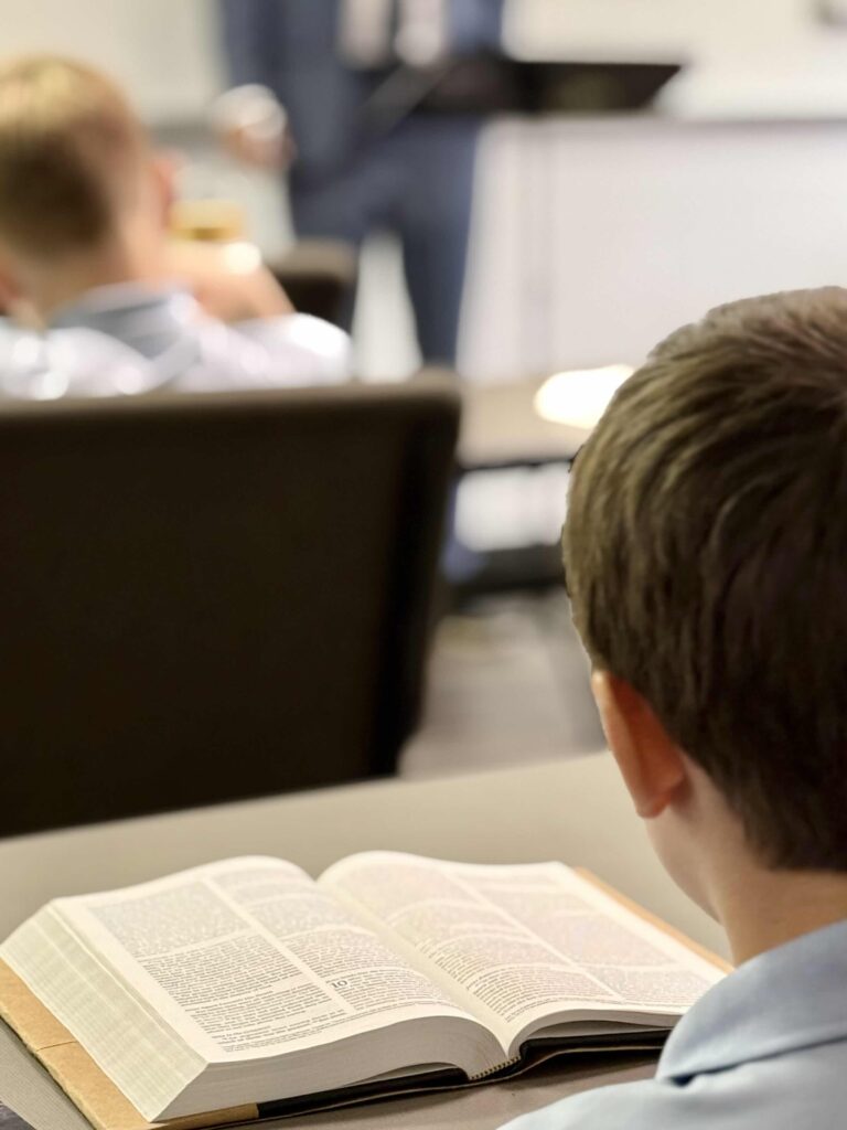 Young boy reads a bible in a classroom setting, with an instructor in the background.