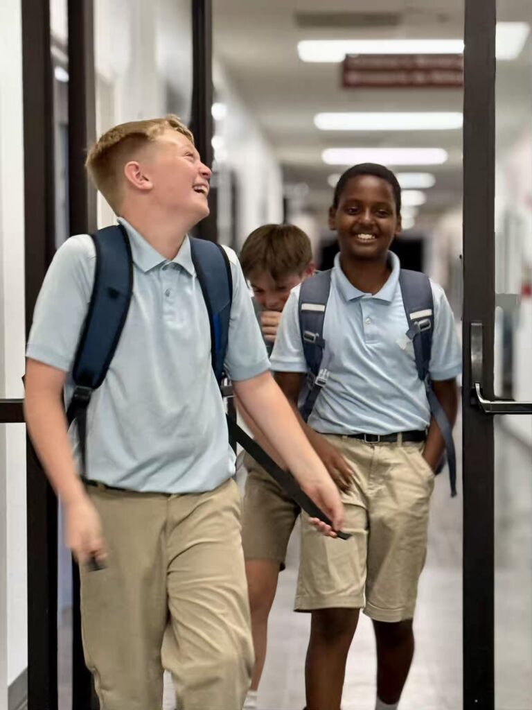 Three smiling school boys in uniform walking down a hallway with backpacks