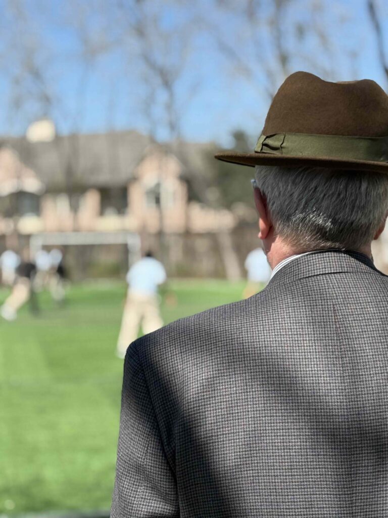 Man in fedora and suit watches soccer game from the sidelines.