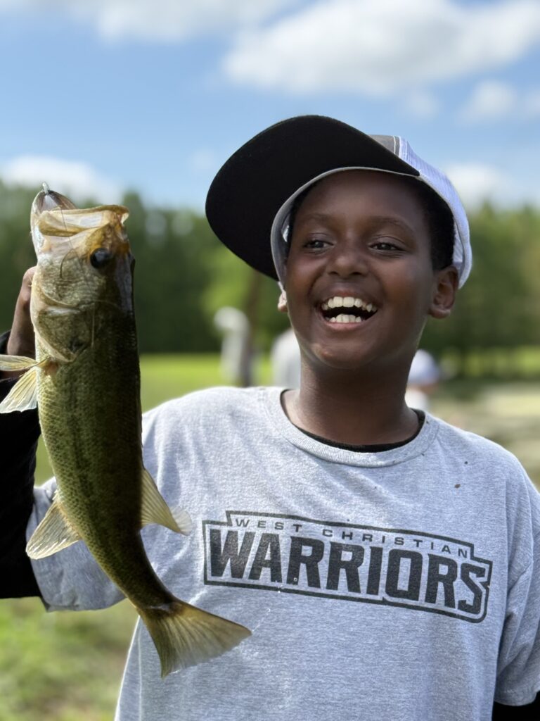 Young boy in a "West Christian Warriors" shirt proudly holds a large bass fish.