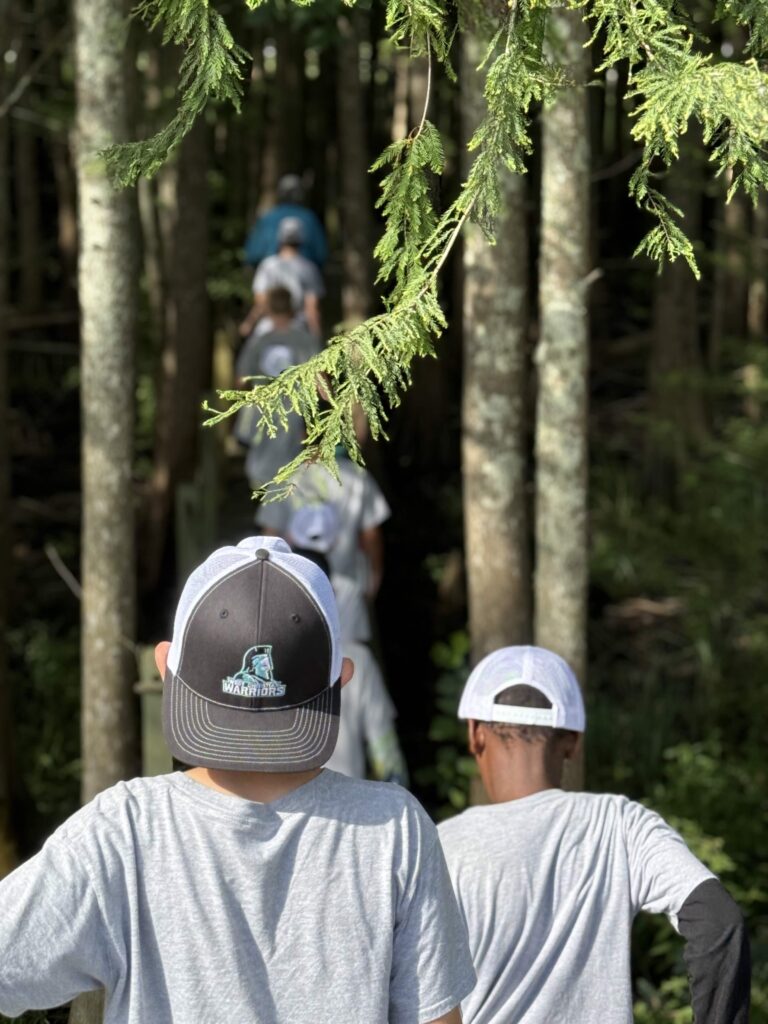 Boys in hats hike on a wooden path through a forest.