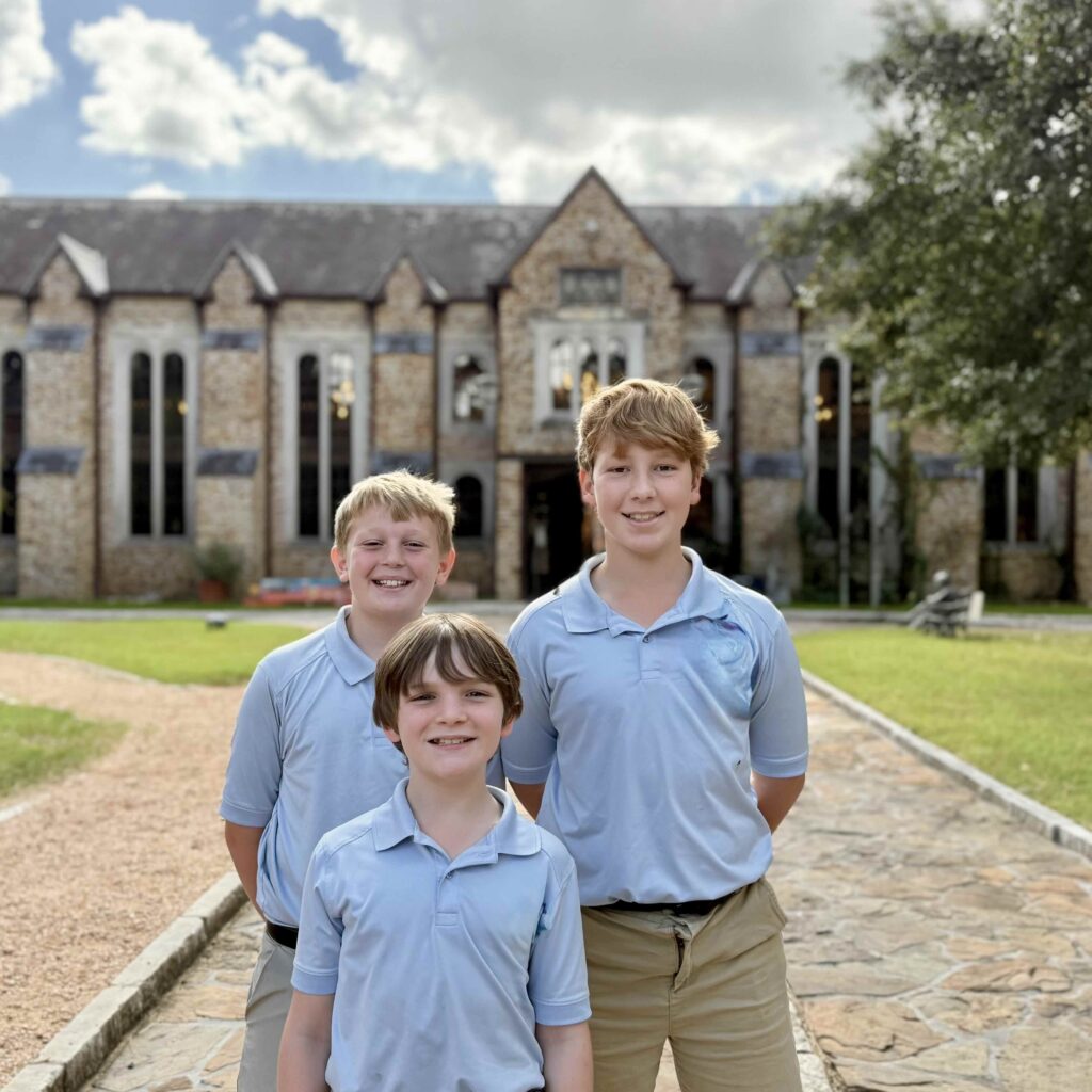 Three smiling boys in light blue polo shirts stand before a stone building.