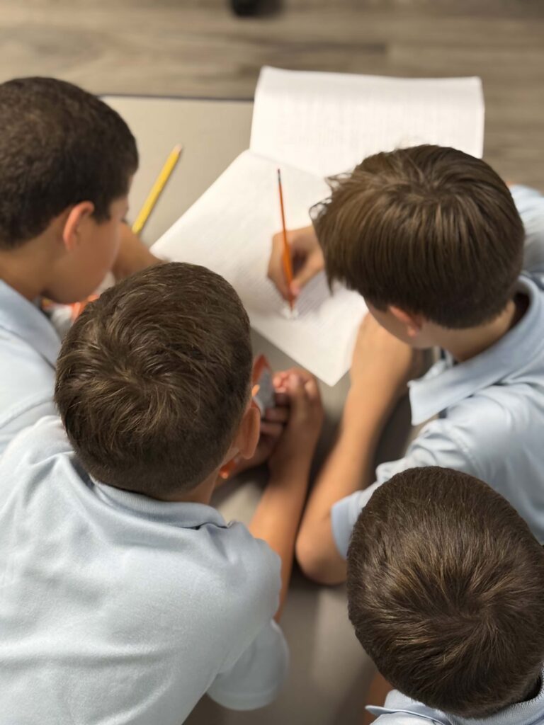 Four young boys in light blue shirts collaborating on a school assignment at a table.