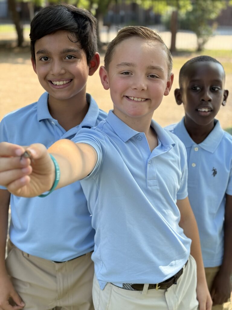 Three young boys in light blue polo shirts smile, one holding up a small object.
